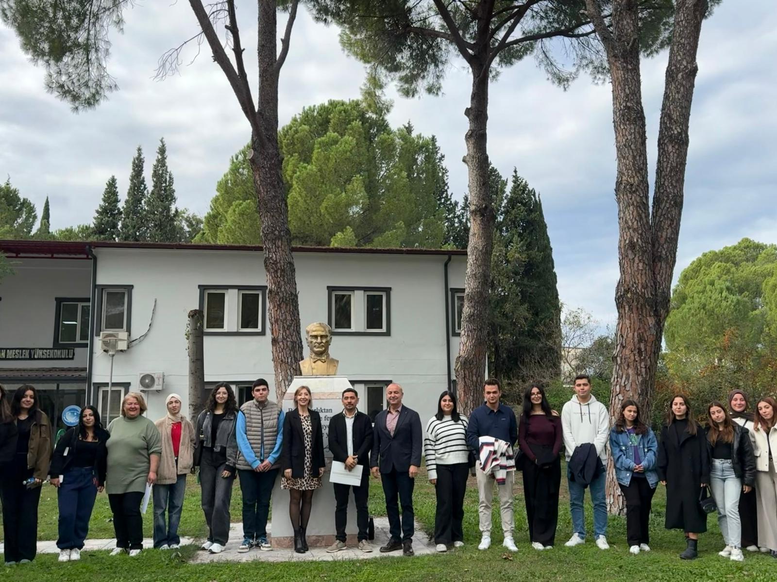 In the photo, a large group of people — likely students along with a few faculty members — are standing outdoors, posing for the camera. Everyone is lined up side by side and smiling. At the center of the group, there is a bust of Atatürk, and behind it stands a white university building. The setting is a green, tree-filled area with tall pine trees creating a natural backdrop. The atmosphere appears friendly yet formal, suggesting that the photo was taken during an academic event. Part of a sign on the building reads “Vocational School.” The sky is slightly cloudy.